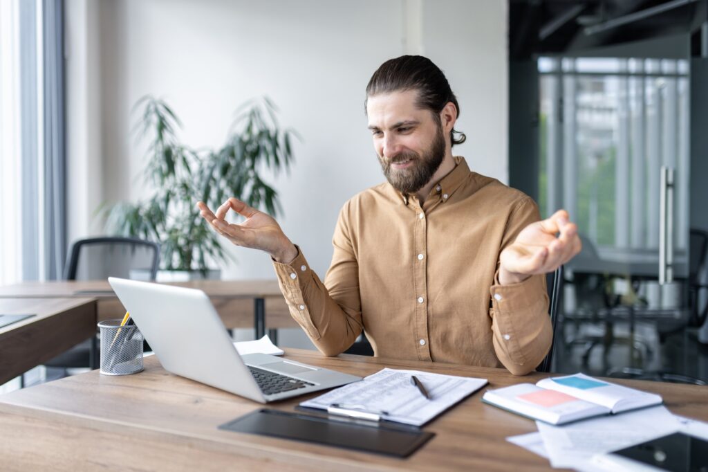 Calm businessman practicing relaxation techniques at work while sitting at desk with laptop and